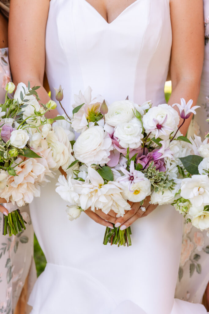 A bride in a white dress holds a bouquet of white and light purple flowers, with other floral bouquets visible nearby—an elegant scene perfectly orchestrated by an Estate Yountville Wedding Planner.