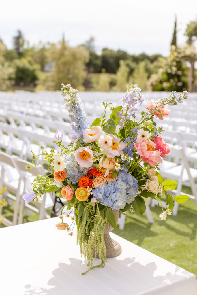 A colorful floral arrangement on a table with rows of white chairs in the background, set outdoors on a sunny day.