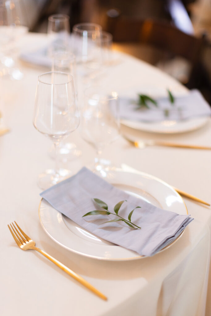 Elegant table setting with white plates, gold cutlery, gray napkins, and a small green branch.