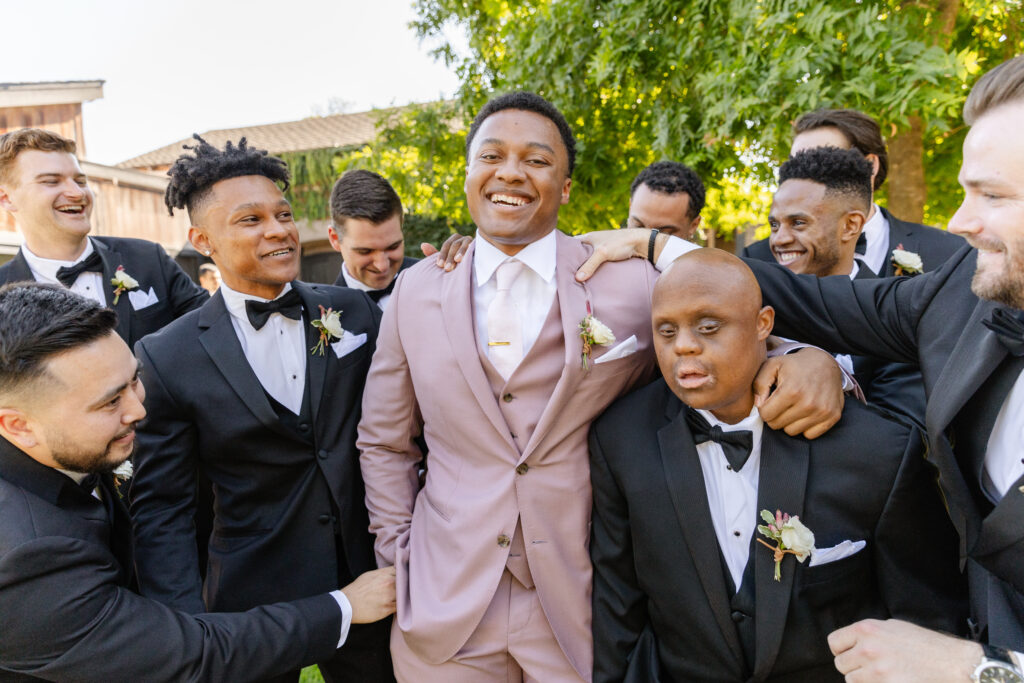 A group of men in tuxedos surround a smiling man in a pink suit outdoors, posing for a photo at what appears to be a formal event expertly coordinated by an Estate Yountville Wedding Planner.