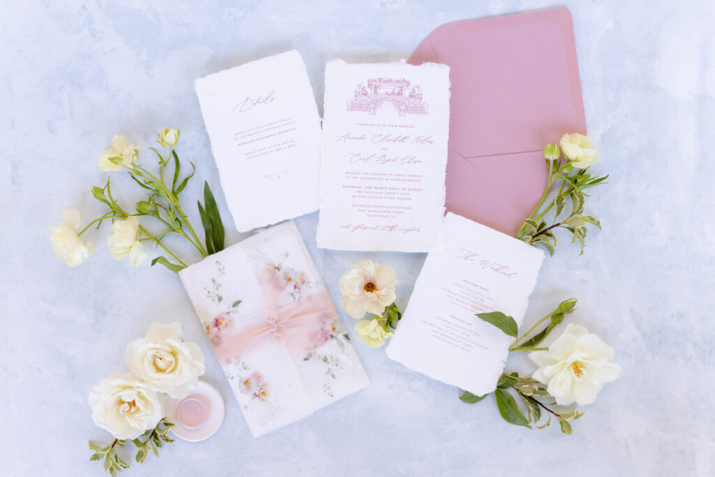 Flat lay of wedding invitations with pink text, a pink envelope, floral decorations, a wax seal, and white roses on a light blue background, perfectly styled by an Estate Yountville Wedding Planner.