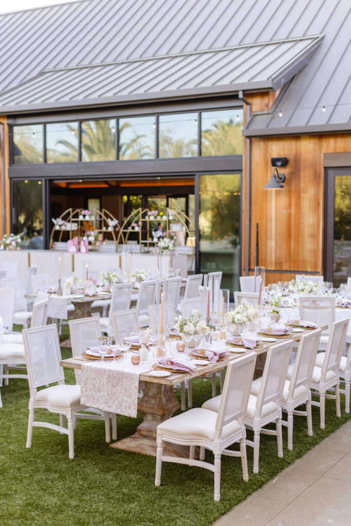 Outdoor wedding reception setup with white chairs, long tables adorned with pink and white floral centerpieces, elegant table runners, and glowing candles, next to a modern wooden building with large windows—beautifully coordinated by an expert Estate Yountville Wedding Planner.