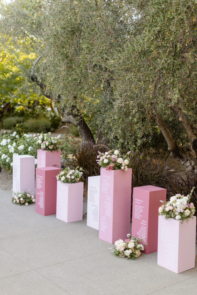 Six pink and white columns display text lists, topped and surrounded by white and blush floral arrangements, set outdoors near trees and greenery on a paved area—an elegant touch curated by an Estate Yountville Wedding Planner.