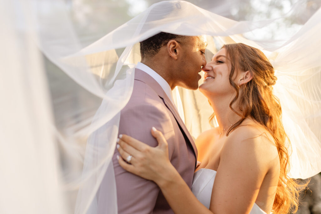 A bride and groom stand close together under a wedding veil, smiling at each other in soft sunlight, their moment perfectly orchestrated by an Estate Yountville Wedding Planner.