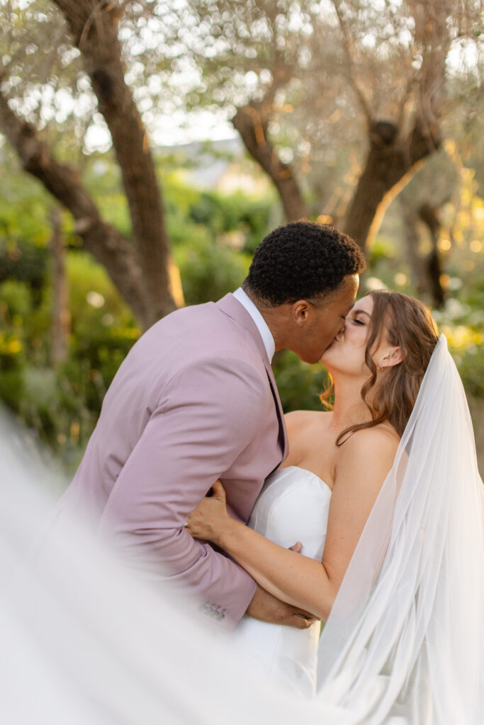 A bride and groom kiss outdoors, surrounded by lush greenery. The groom wears a light mauve suit, and the bride is in a strapless white dress with a long veil—an elegant moment beautifully orchestrated by an Estate Yountville Wedding Planner.