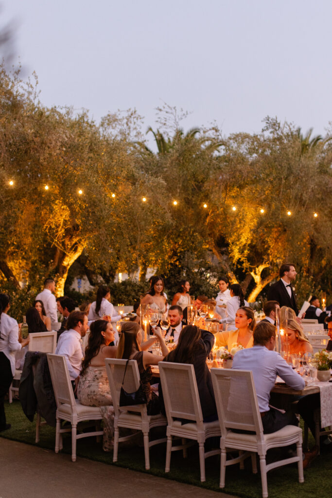 A group of people sit at a long outdoor table with string lights and candles, dining and talking in a garden setting during the evening—an enchanting ambiance that evokes the touch of an Estate Yountville Wedding Planner.