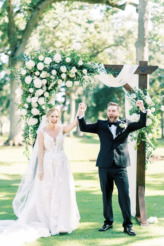 A bride and groom celebrate under a floral arch outdoors. The bride wears a white dress and veil, and the groom is in a black suit, holding hands and smiling.