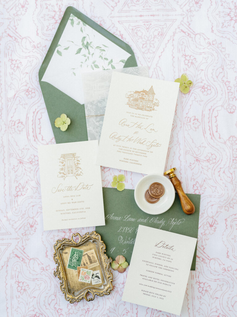 Wedding invitation suite with green and white stationery, ornate envelope, stamps, wax seal, and decorative dish on a patterned fabric.