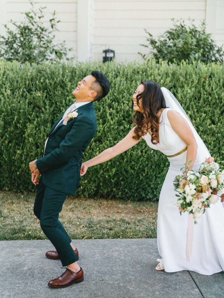 A bride playfully tugs at the groom's jacket, both laughing, in front of greenery. She holds a bouquet of flowers.