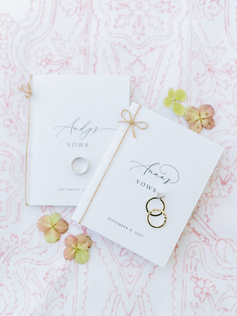 Two vow booklets with gold rings on one and a silver ring on the other, tied with twine, on a pink patterned fabric with scattered hydrangea petals.