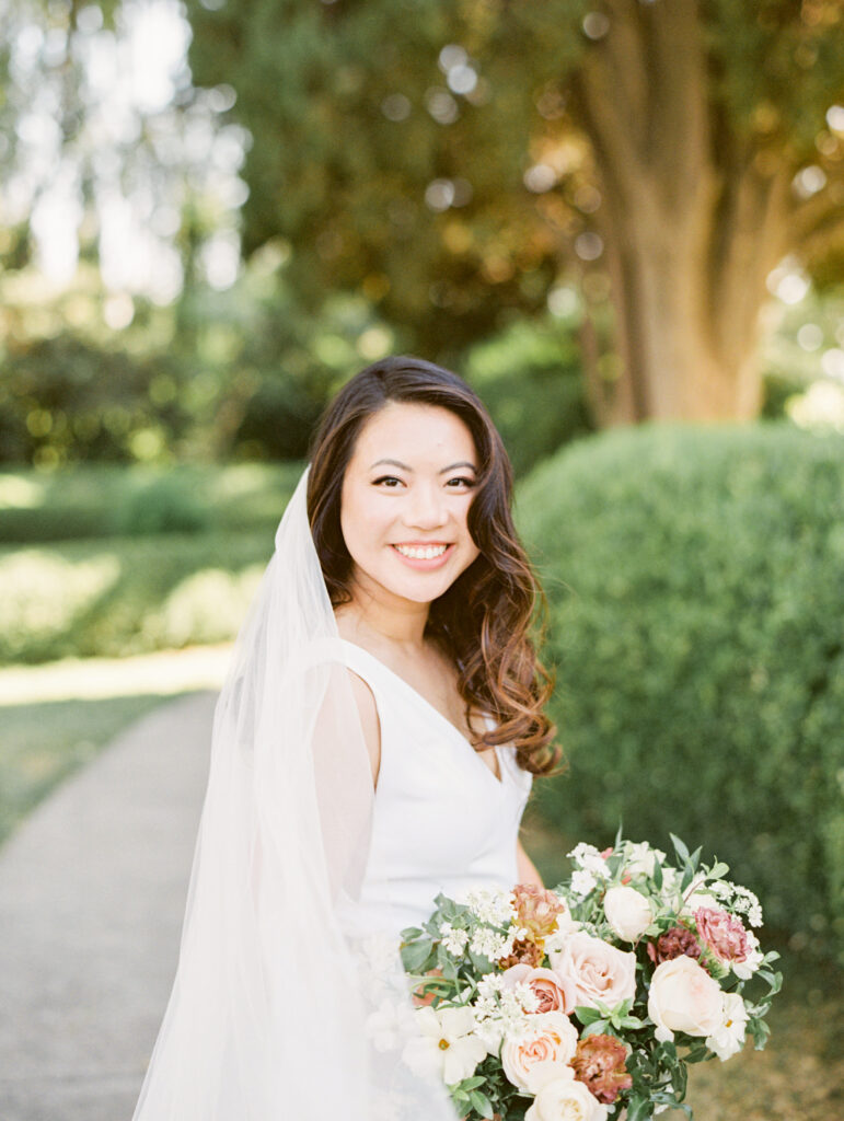 A bride in a white dress smiles while holding a bouquet of flowers. She is standing outdoors with greenery in the background.