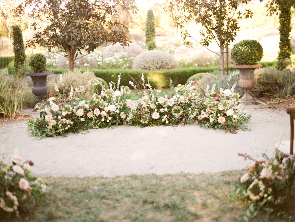 A circular arrangement of flowers and greenery in a garden setting with manicured bushes and trees, bathed in soft sunlight.