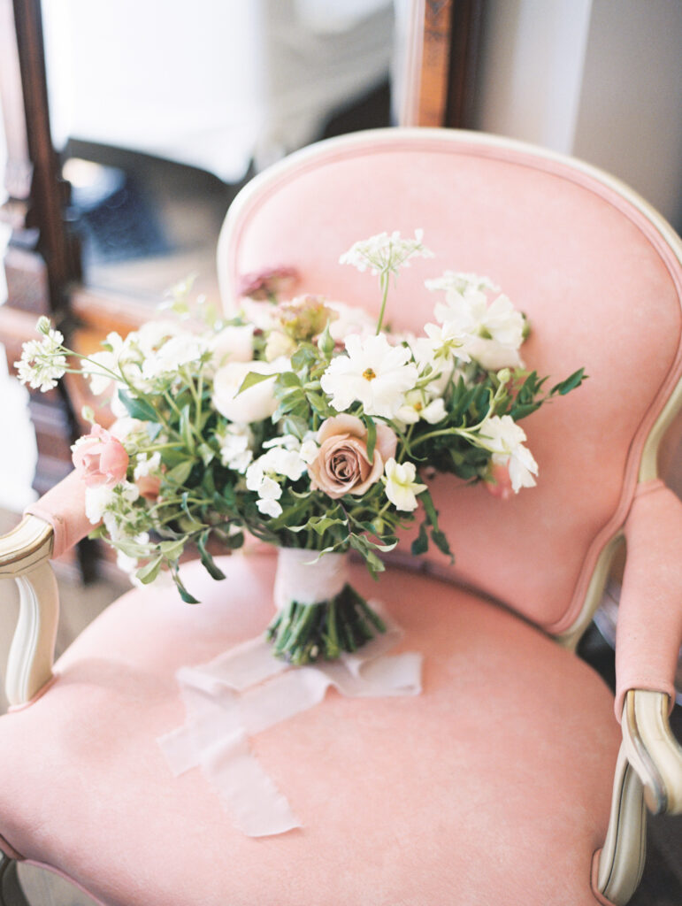 A bouquet of flowers with roses and greenery rests on a pale pink upholstered chair.