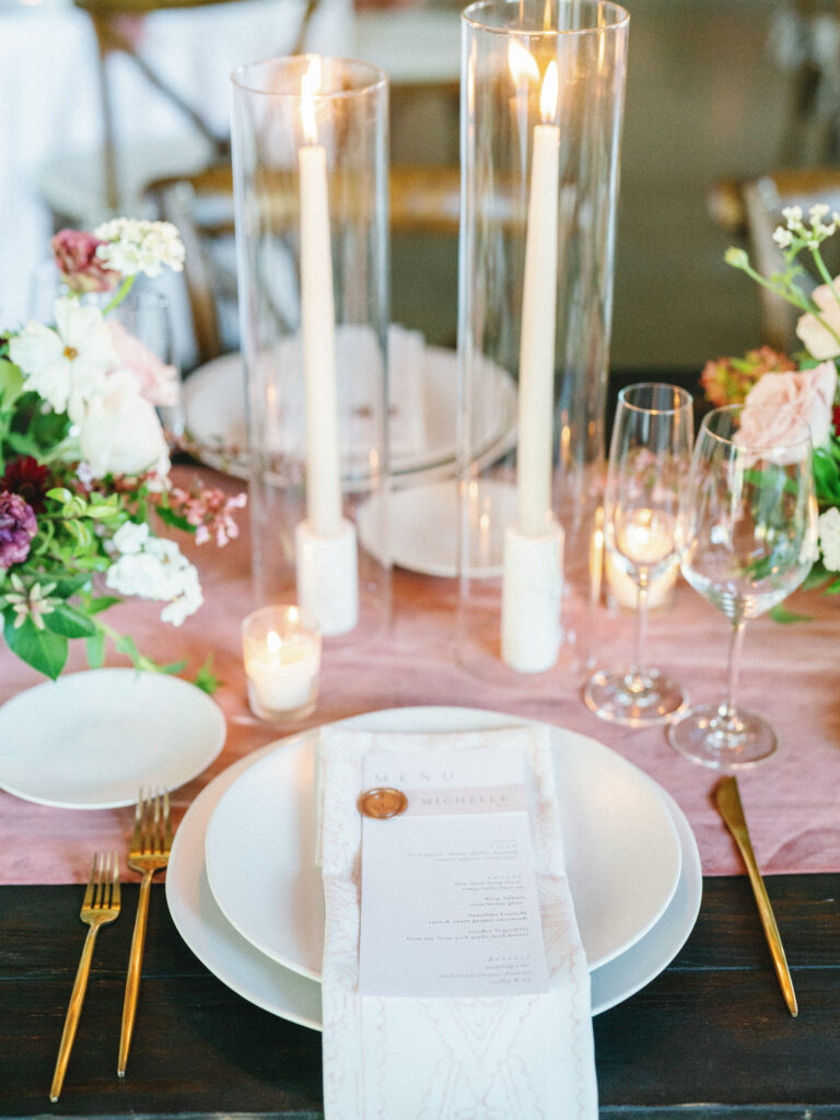 Elegant table setting with white plates, a menu, gold cutlery, and glasses. Tall candles in glass holders and floral arrangements decorate the table on a pink runner.