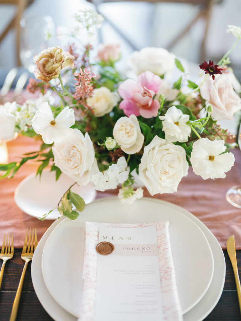 A beautifully arranged dining table with an elegant floral centerpiece, featuring white and pink flowers. A menu is placed on a plate, accompanied by gold cutlery.