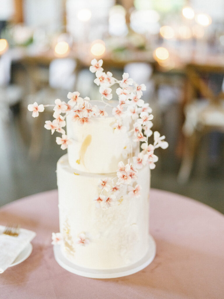 A two-tiered white cake decorated with delicate pink cherry blossoms sits on a pink tablecloth.