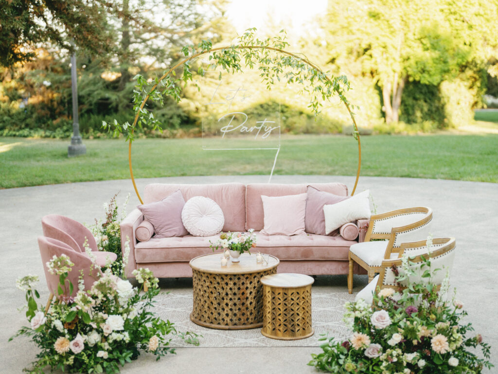 Outdoor seating area with a pink sofa, chairs, and floral arrangements, featuring a circular frame with greenery.