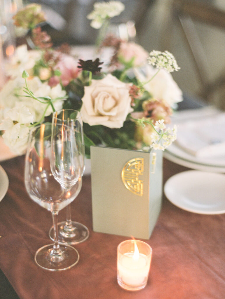 A table setting with a floral arrangement in a box, two empty wine glasses, a small lit candle, and a white plate.