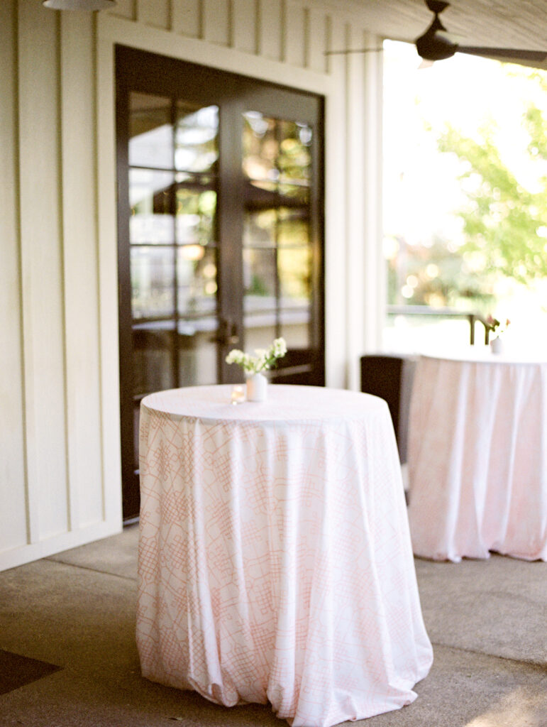 Round table with a white cloth and small flower vase on a patio. Another similar table is in the background near a glass door.