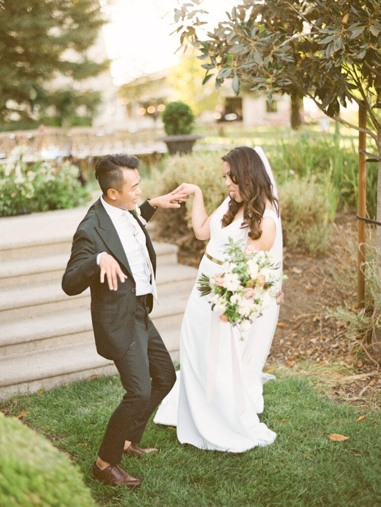 A groom and bride in a playful pose outdoors, with the bride holding a bouquet and both dressed in formal attire.