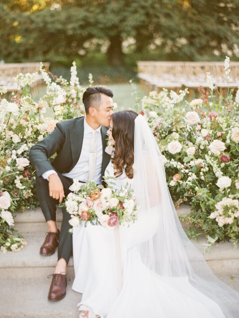A bride and groom sit on steps surrounded by flowers. The groom is in a suit, and the bride wears a veil, holding a bouquet. They lean closely together.