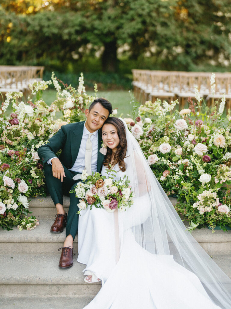 A couple in wedding attire sits on steps surrounded by lush floral arrangements and greenery.