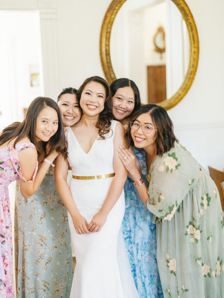A bride in a white dress stands smiling with four bridesmaids in floral dresses, posing in front of a large round mirror.