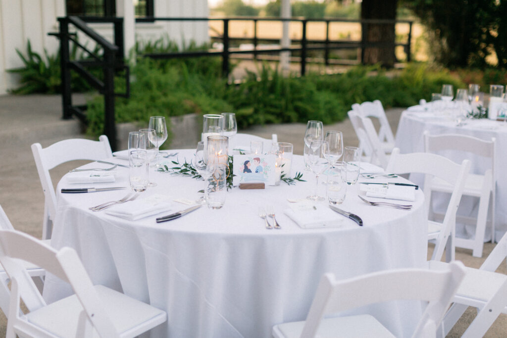 A round table with a white tablecloth is set with glassware, napkins, and candles. White chairs surround the table, and greenery decorates the center. The setting is outdoors.