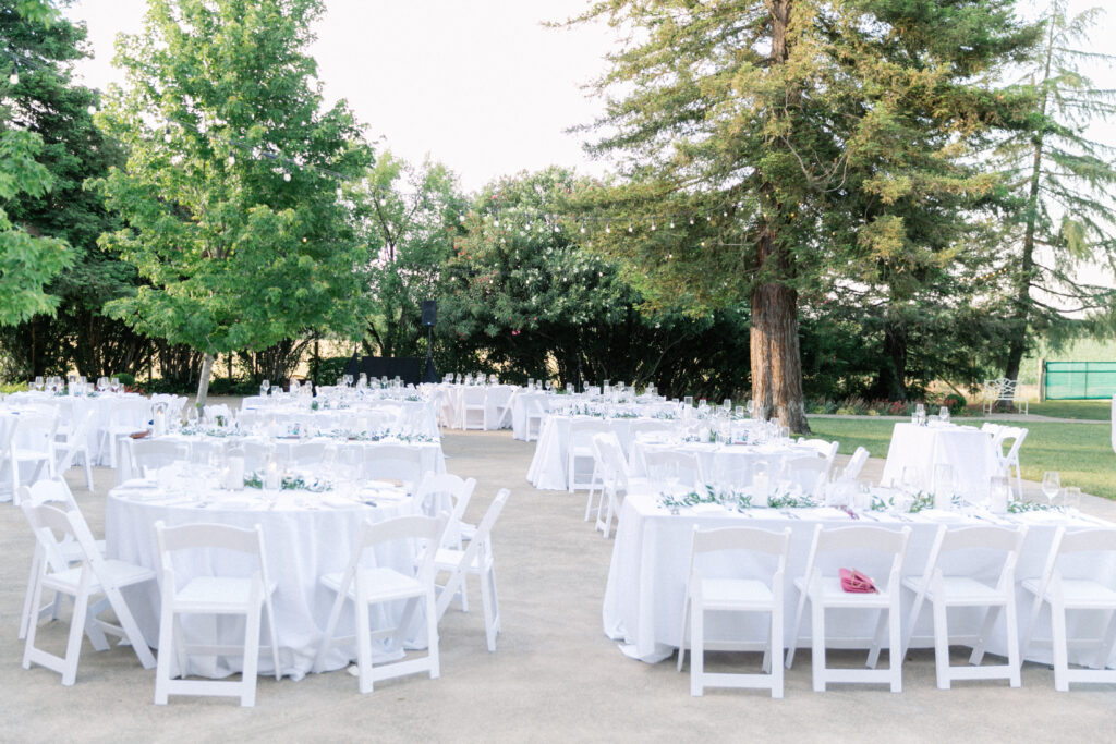 Outdoor event setup with multiple round tables covered in white tablecloths and surrounded by white folding chairs, under large trees.