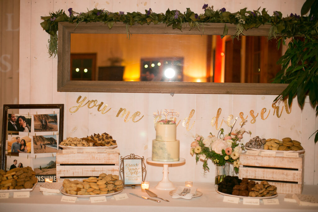 A dessert table with a cake, various cookies, and photos in frames. A sign reads "you, me and dessert" and there are flowers and small lit candles for decoration.