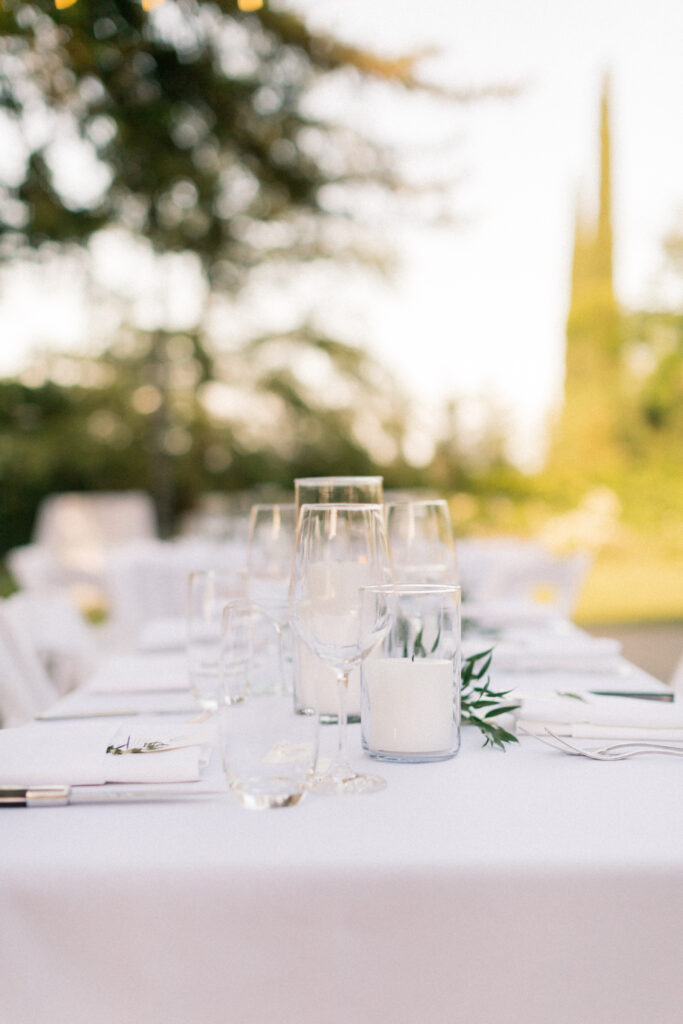 Outdoor table setting with white tablecloth, multiple empty glasses, and a blurred background of trees and greenery.