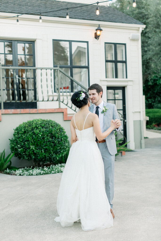 A couple dances outside, the woman in a white dress and the man in a gray suit, in front of a house with string lights.