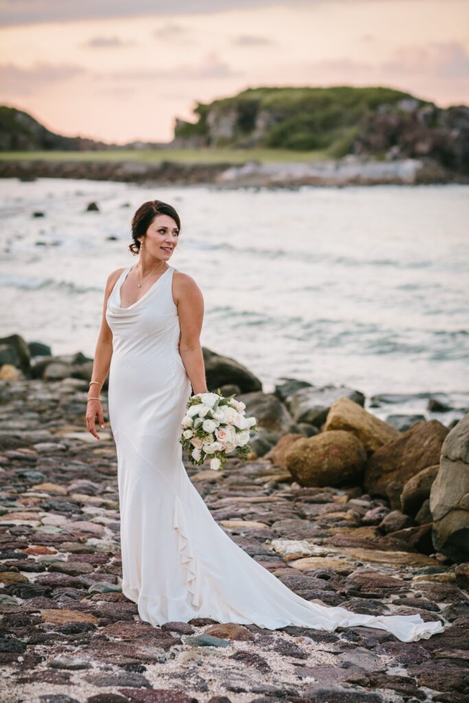 A woman in a white wedding dress stands on a rocky beach holding a bouquet, with the ocean and a grassy cliff in the background.