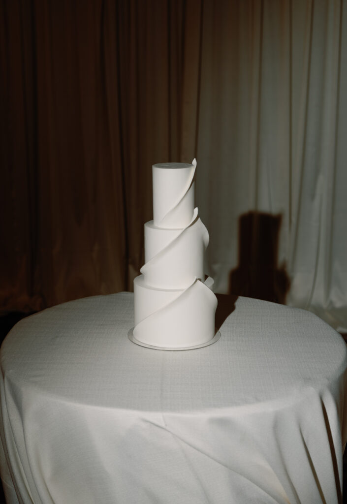 Three-tiered white wedding cake with smooth, spiral design on a round table covered with a white tablecloth.