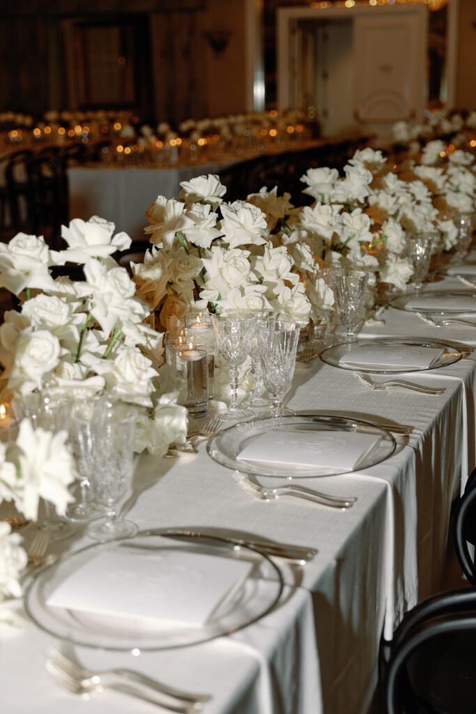 A table set for an event with white tablecloth, glassware, plates, and white floral arrangements in vases.