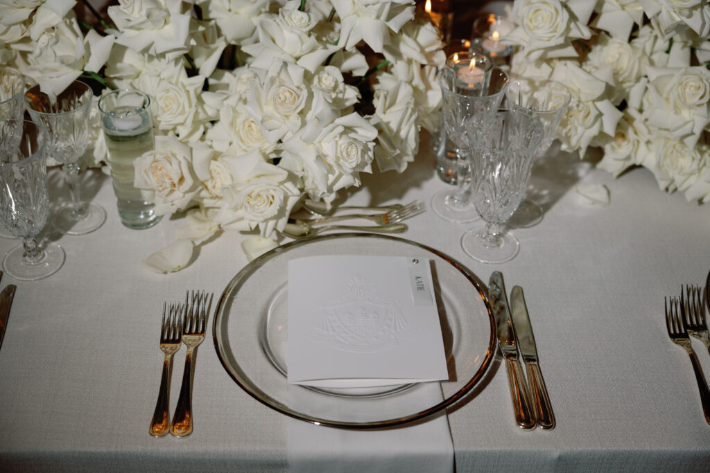 A formal dining table setting with white roses, crystal glassware, a place card, and silver cutlery on a white tablecloth.