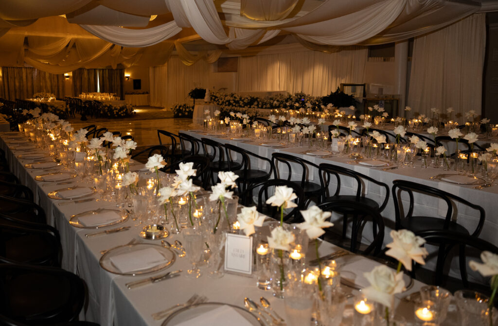 Elegant banquet hall with long tables set for an event. White flowers, candles, and black chairs adorn the setting, under draped fabric ceiling.