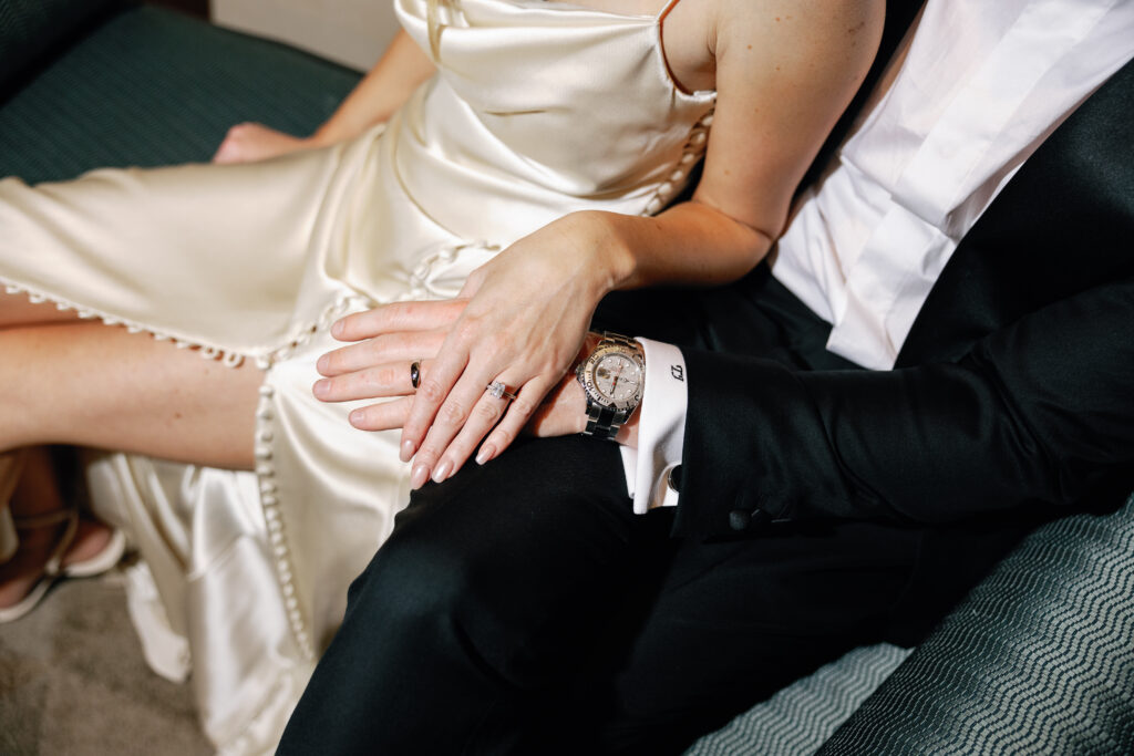 A couple in formal attire sit closely on a couch. They gently hold hands, showcasing their rings. The focus is on their hands and clothing details.