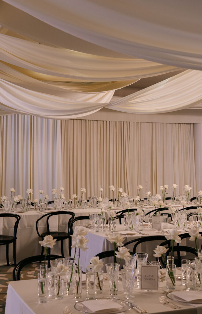 Elegant banquet hall with white draped ceiling, set tables with white flowers, glassware, and black chairs.