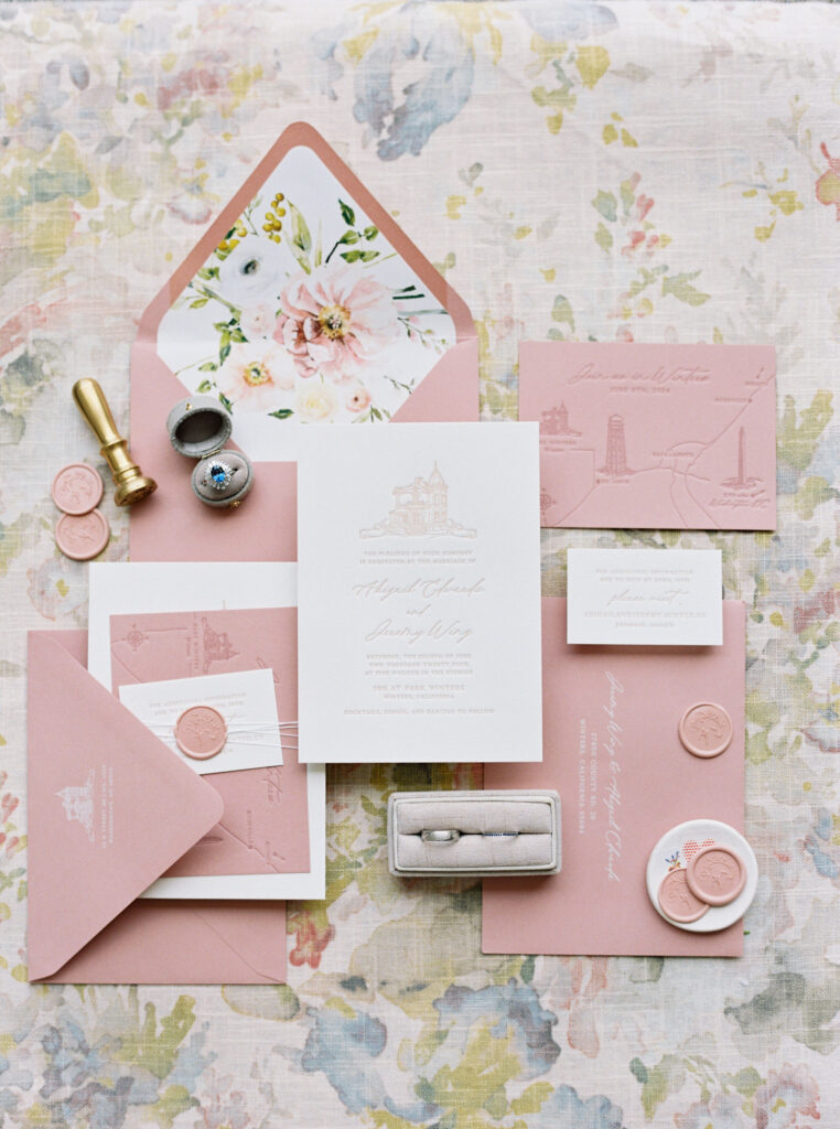 A floral-themed wedding invitation set with pink envelopes, invitation, cards, wax seals, a ring, stamp, and pen displayed on a floral patterned cloth background.