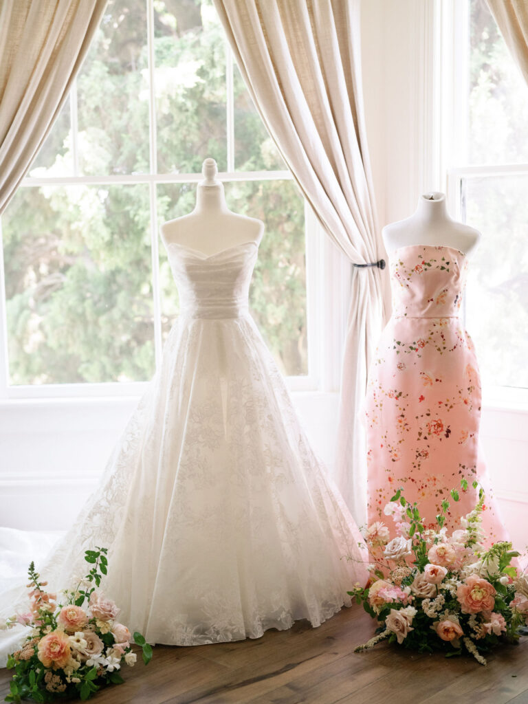 Two mannequins display elegant gowns, one white and one pink, surrounded by floral arrangements in front of large windows with light curtains.