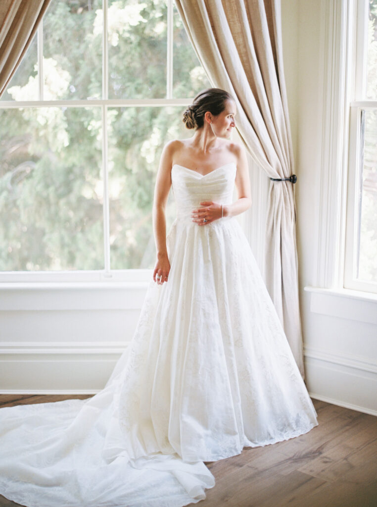 Bride in a white wedding gown stands by a large window with light curtains, looking to the side.