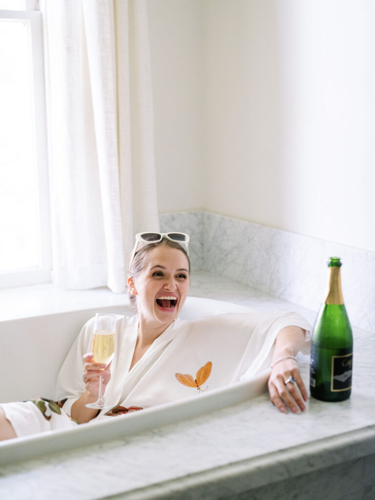 A woman in a bathrobe relaxes in a marble bathtub, holding a glass of champagne, with a bottle nearby, smiling broadly.