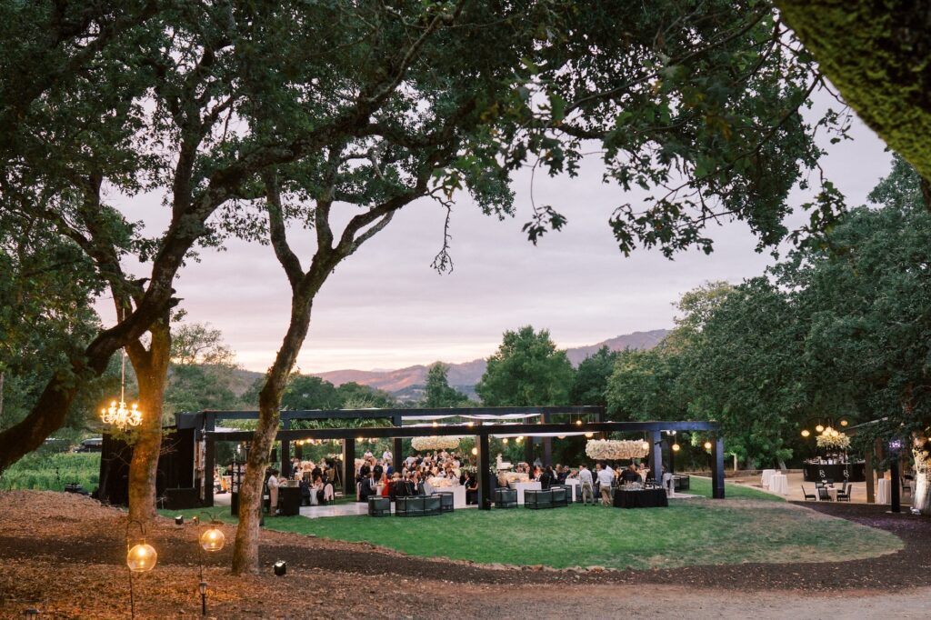 Outdoor event under large trees with people gathered around tables. Elegant lighting and decorations are visible, set against a backdrop of hills during sunset.