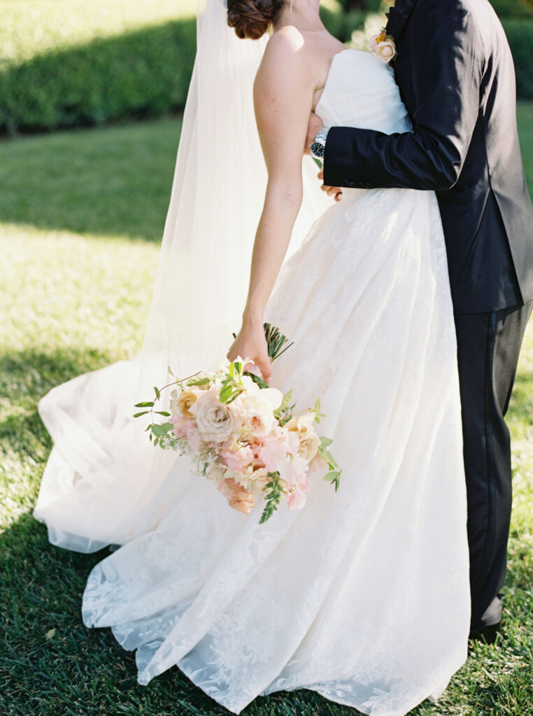 A bride and groom embrace outdoors. The bride holds a bouquet of flowers and wears a strapless white gown and veil. The groom is in a dark suit.
