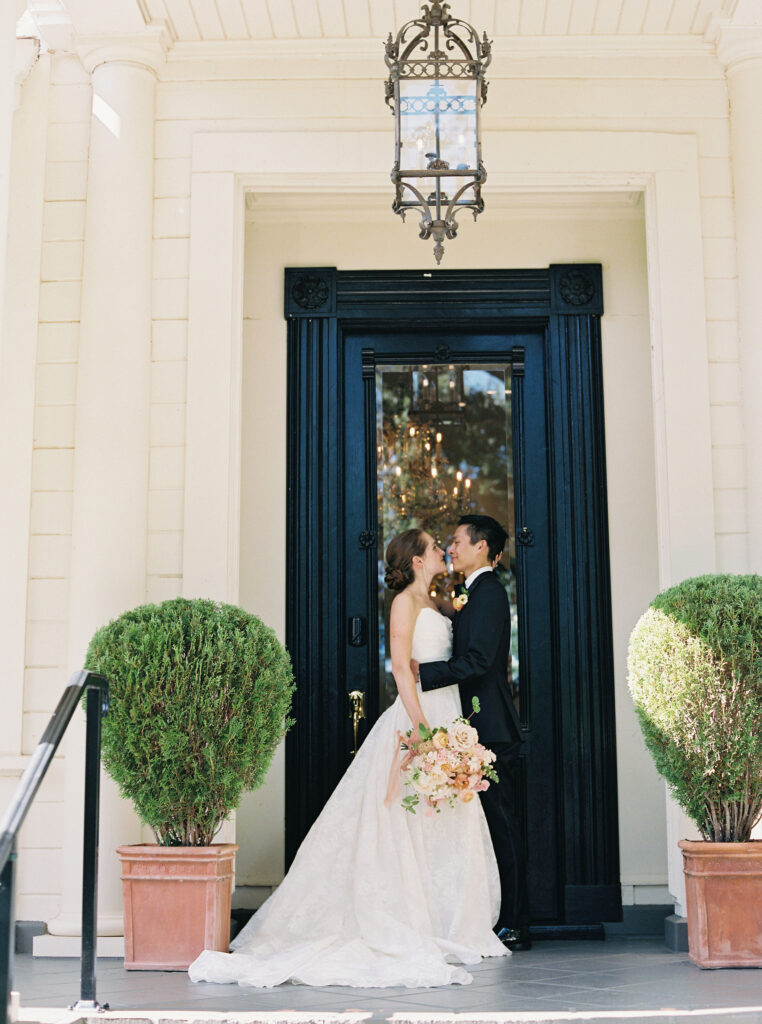 A bride and groom stand under a lantern at the entrance of a building, surrounded by potted plants. The bride holds a bouquet while they face each other.