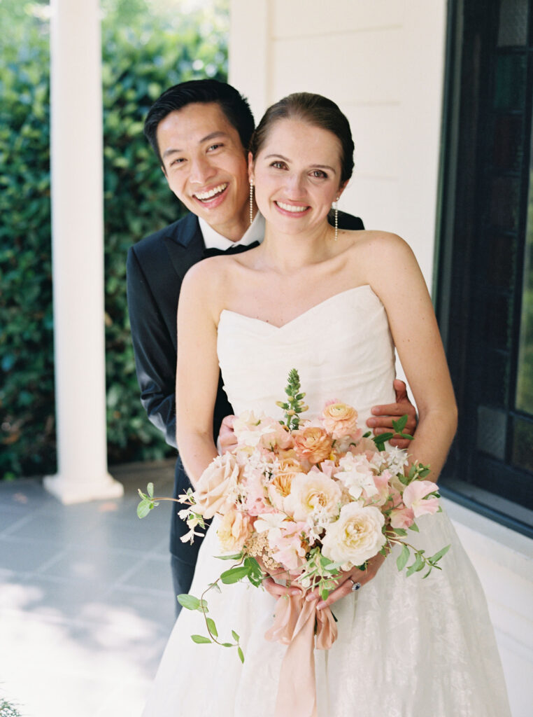 A couple in wedding attire smiles, with the bride holding a bouquet of pink and white flowers, standing on a porch.