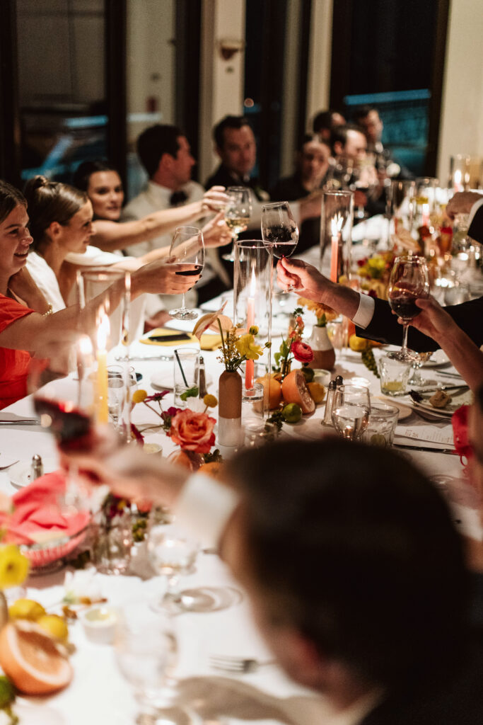 A group of people seated at a long dining table raise their glasses for a toast. The table is decorated with flowers and candles.