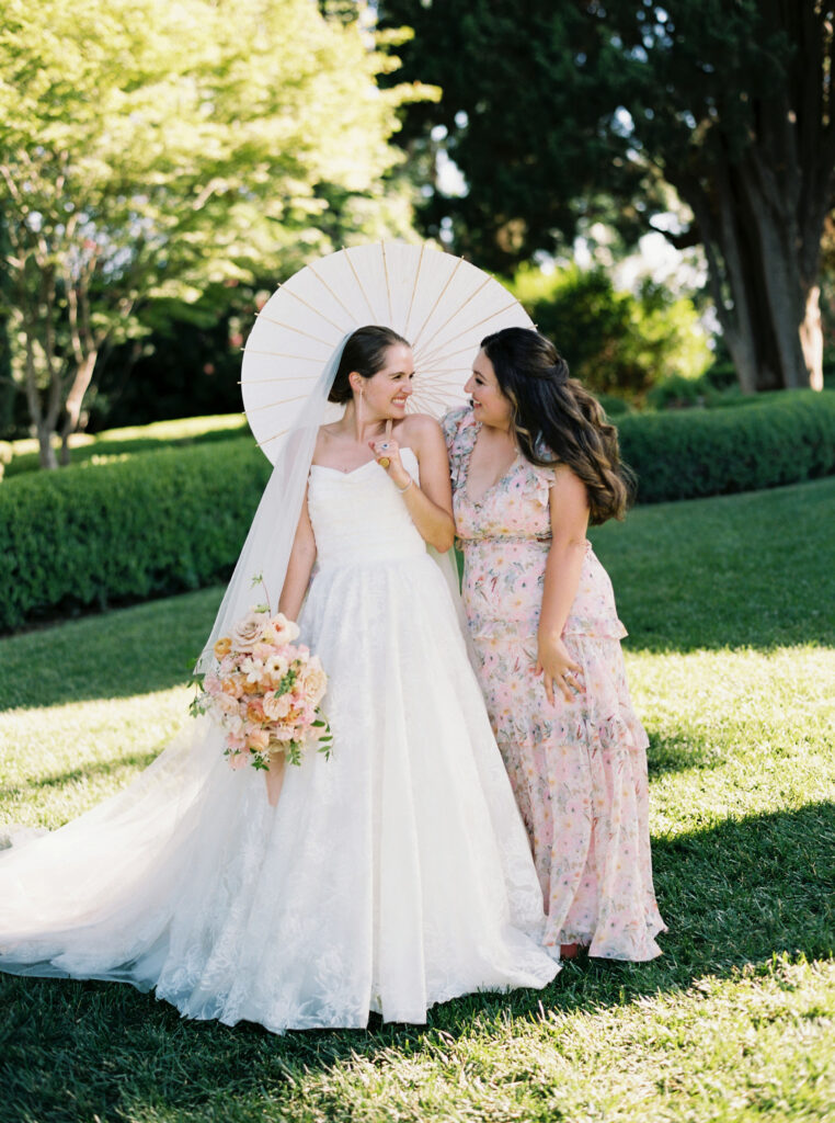 Bride in a white dress holding a parasol stands with a woman in a floral dress, both smiling in a garden setting.