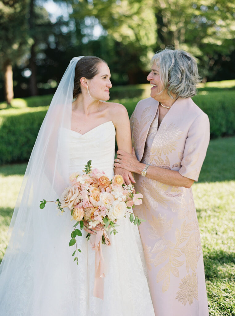 A bride in a white dress holding a bouquet stands next to a woman in a light pink dress outdoors, both smiling at each other.